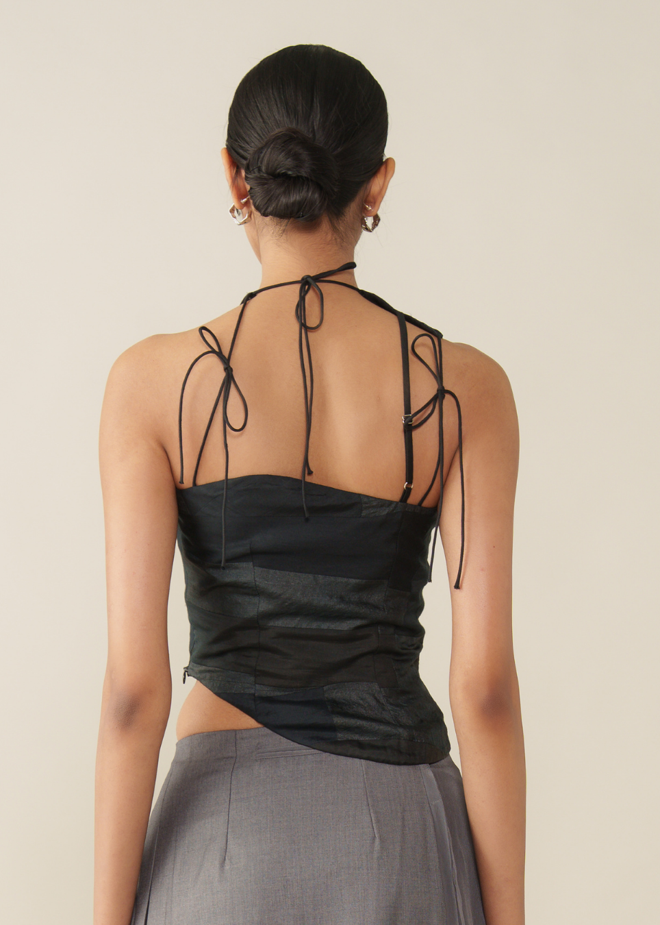 A woman shown from the back wearing a black halter-style top with tie straps at the shoulders and a gathered waist, paired with a gray skirt, against a plain light beige background.