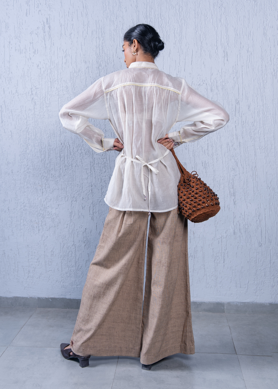 A woman stands with her back to the camera wearing a sheer white long-sleeve blouse with a tie waist and wide-leg beige trousers, holding a brown woven handbag against a textured light gray wall.