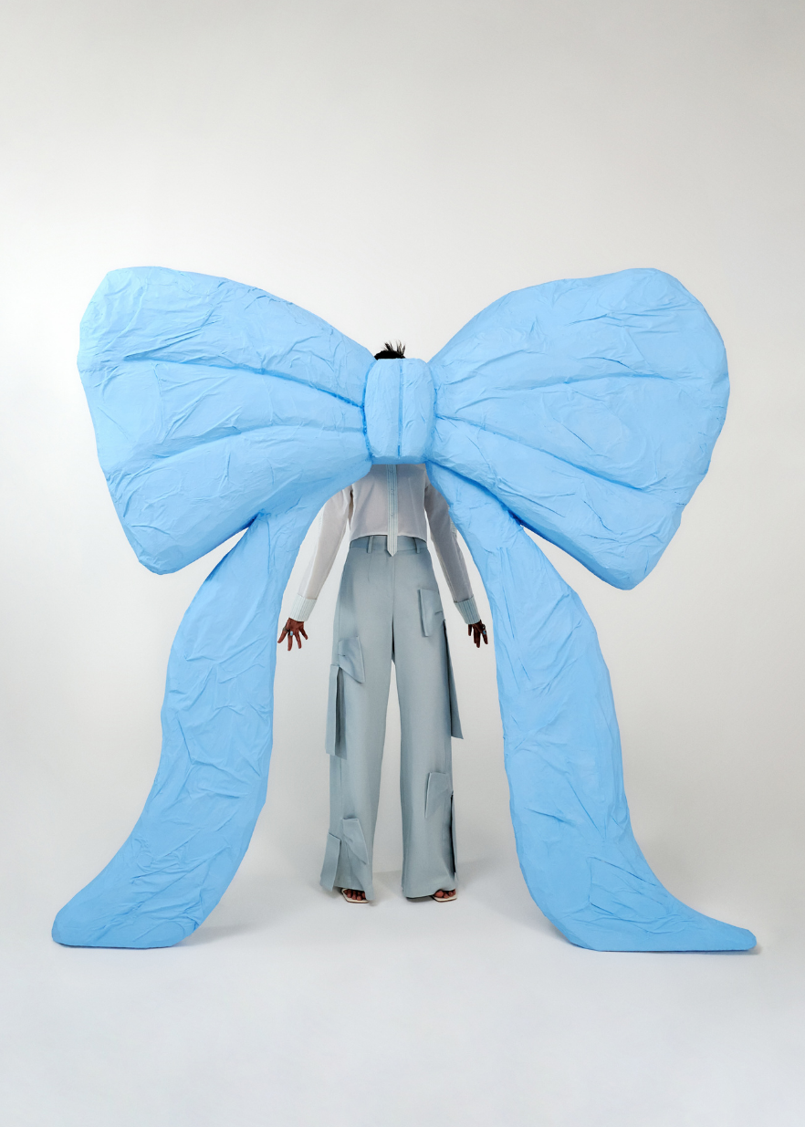 A person stands facing a giant light blue bow-shaped backdrop or costume in a studio setting. The person is wearing a white top and light blue wide-leg pants.