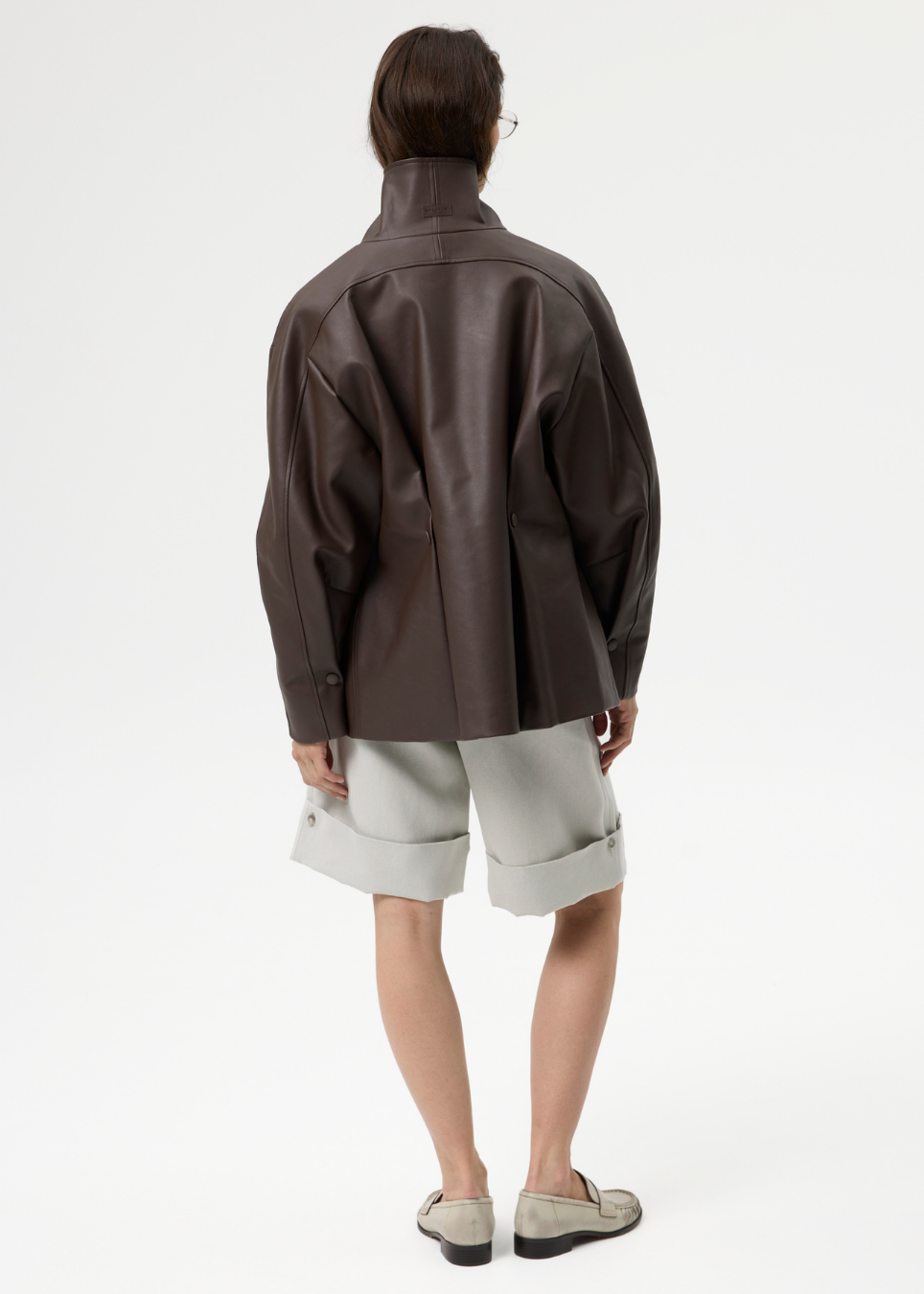 A model shown from the back wearing a dark brown leather jacket, light gray rolled-up shorts, and gray slip-on shoes against a plain white studio background.