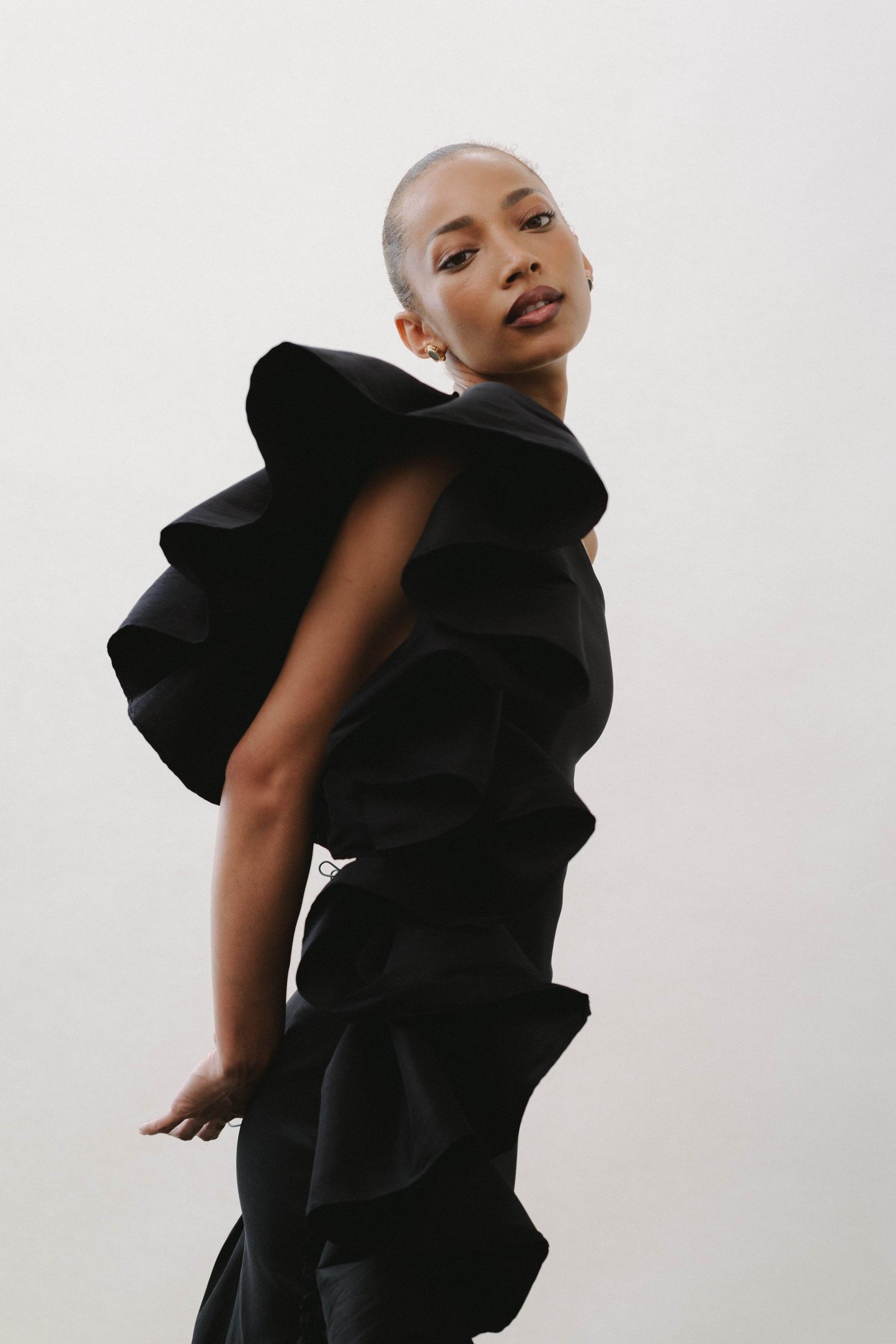 A model wearing a black asymmetrical tight-to-the-body taffeta dress with volume detail on the left side, standing against a white background.