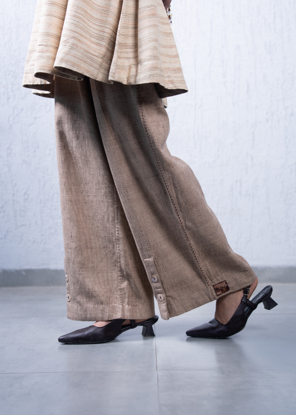 A person wearing wide-leg taupe linen trousers with button details and a beige striped top, paired with black slingback kitten heels, standing against a textured light gray wall.