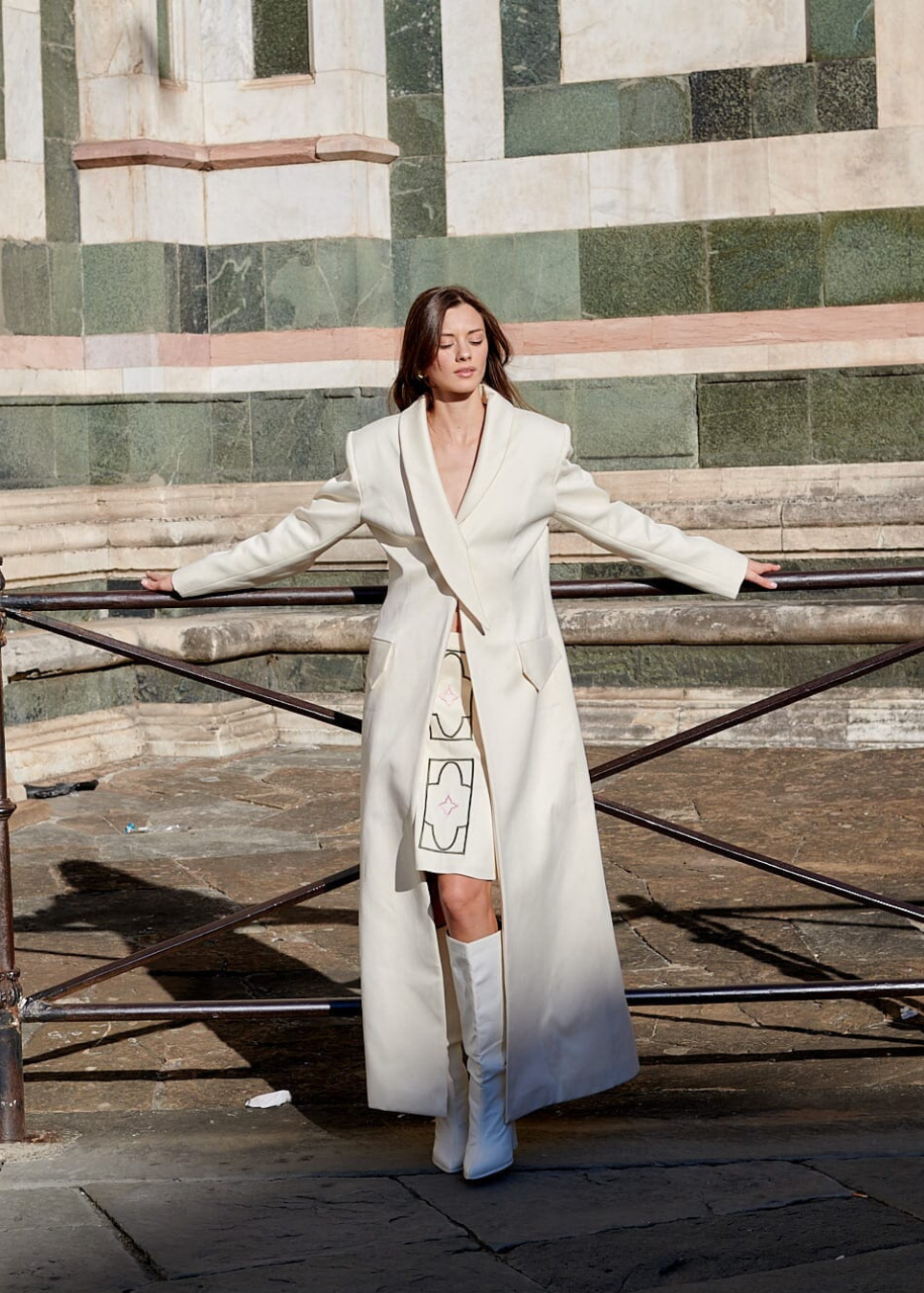 A woman stands outdoors in front of a stone building and metal railing, wearing a long cream coat with a front flap detail and matching cream knee-high boots.