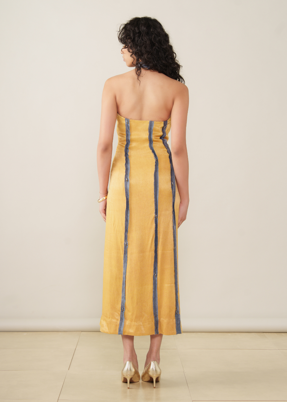 A woman shown from the back wearing a strapless yellow midi dress with vertical blue stripes, paired with metallic gold heels, set against a plain beige studio background.