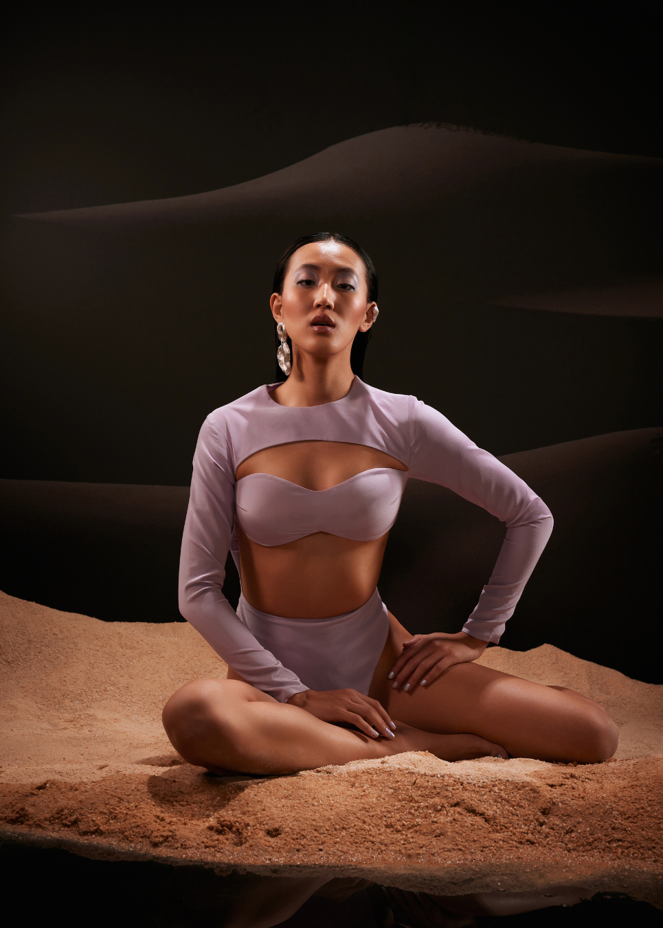 Woman in a purple bodysuit sitting on sand with a dark background