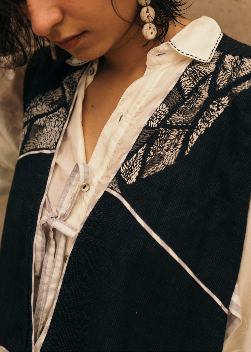 Close-up of a person wearing a dark navy textured vest over a white collared shirt, with visible white contrast stitching and a geometric leaf-like pattern across the shoulders.