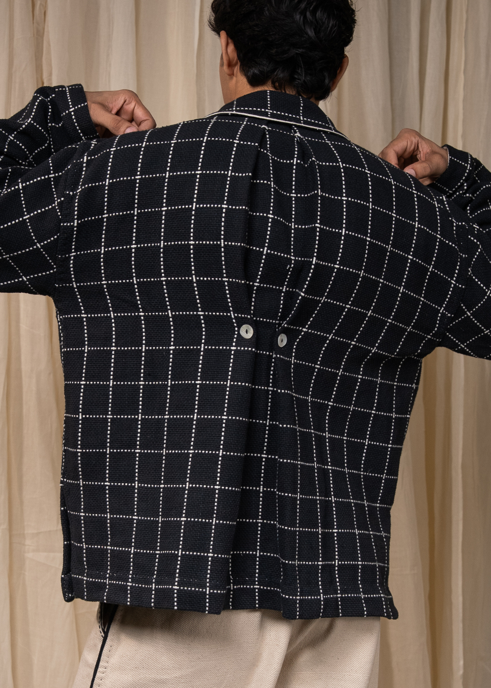 A man viewed from behind wearing a black overshirt with a white dotted windowpane grid pattern, paired with beige trousers, set against a neutral beige curtain background.