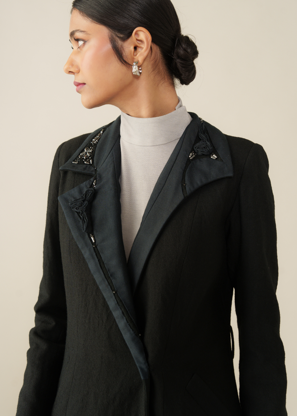 A woman in profile wearing a black tailored jacket with dark satin lapels and decorative beaded embellishments on the shoulders, layered over a light gray turtleneck.