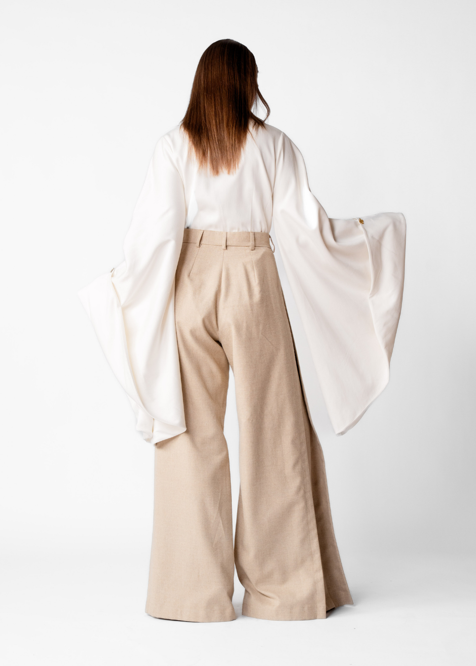 A woman shown from the back wearing a white long-sleeve blouse with dramatic wide sleeves and beige wide-leg trousers, photographed against a clean light gray studio background.