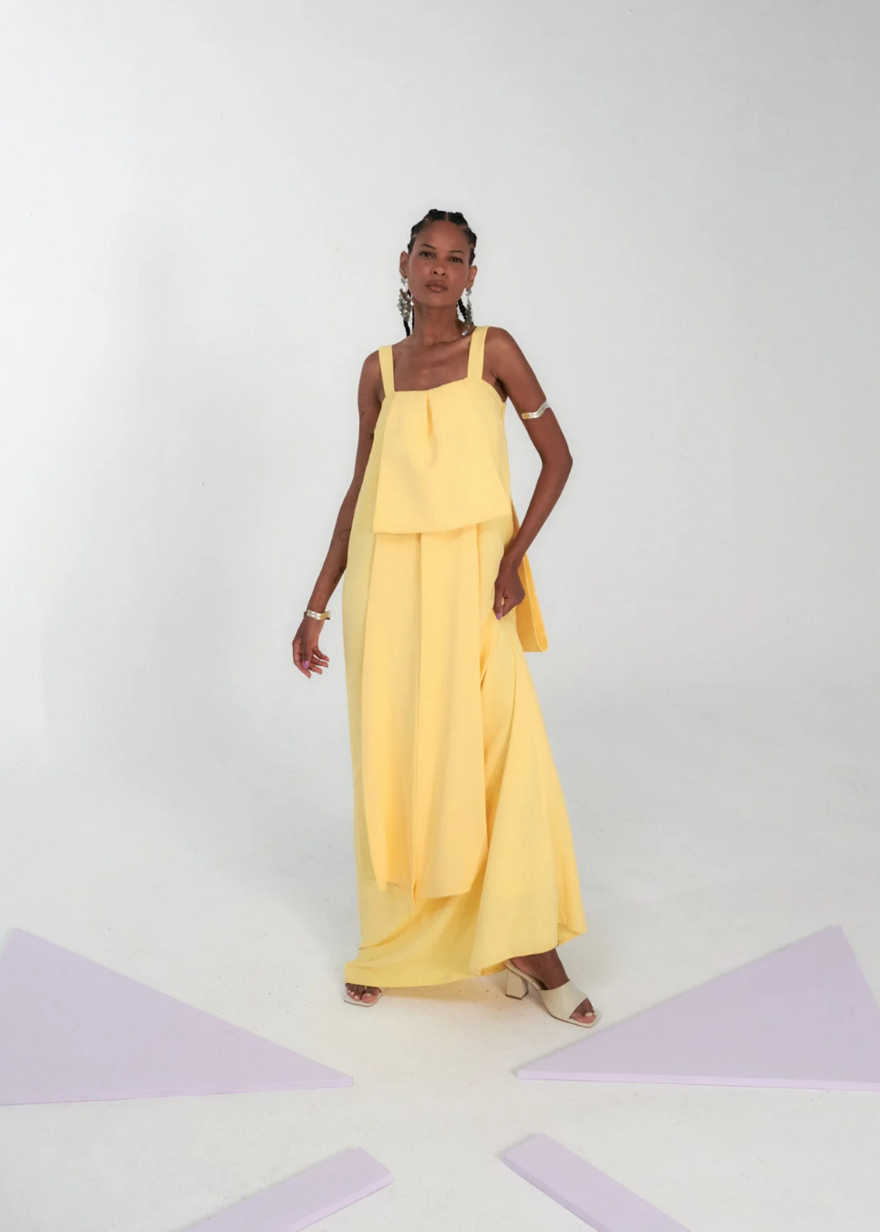 A model wearing a pale yellow sleeveless maxi dress with a draped, layered design and wide-leg pants, standing against a white studio background with light purple geometric shapes on the floor.