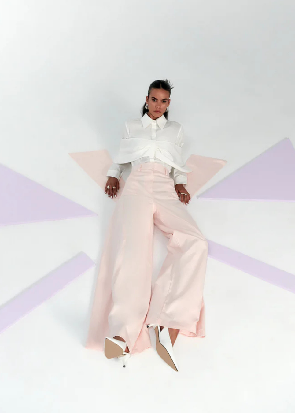 A model wearing a white long-sleeve blouse with dramatic ruffled shoulders and high-waisted, wide-leg blush pink trousers, posed on a white floor with light purple geometric shapes.