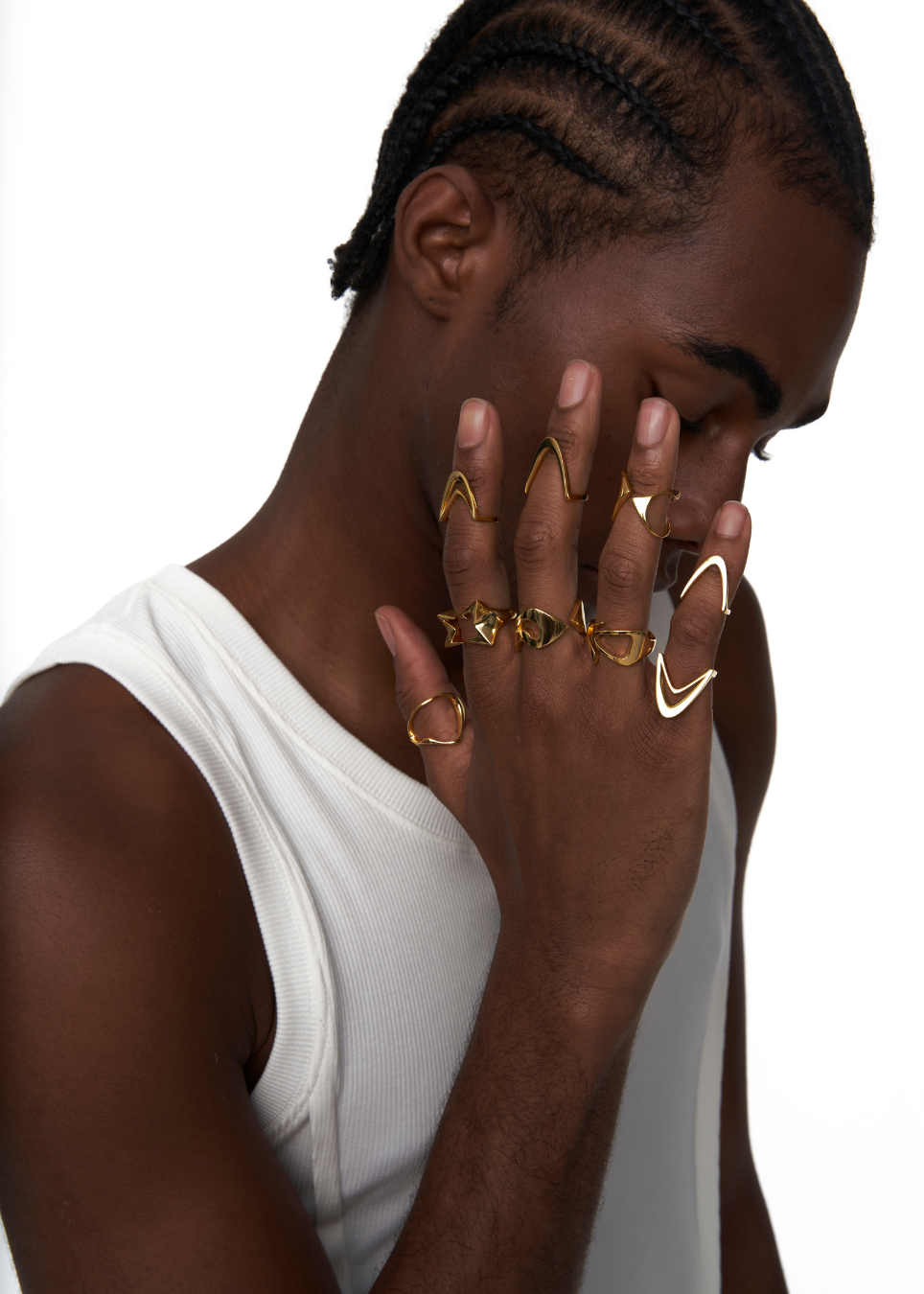 A close-up of a man wearing multiple gold rings on his fingers, including a large geometric ring on his middle finger and a curved ring on his ring finger, against a white background.