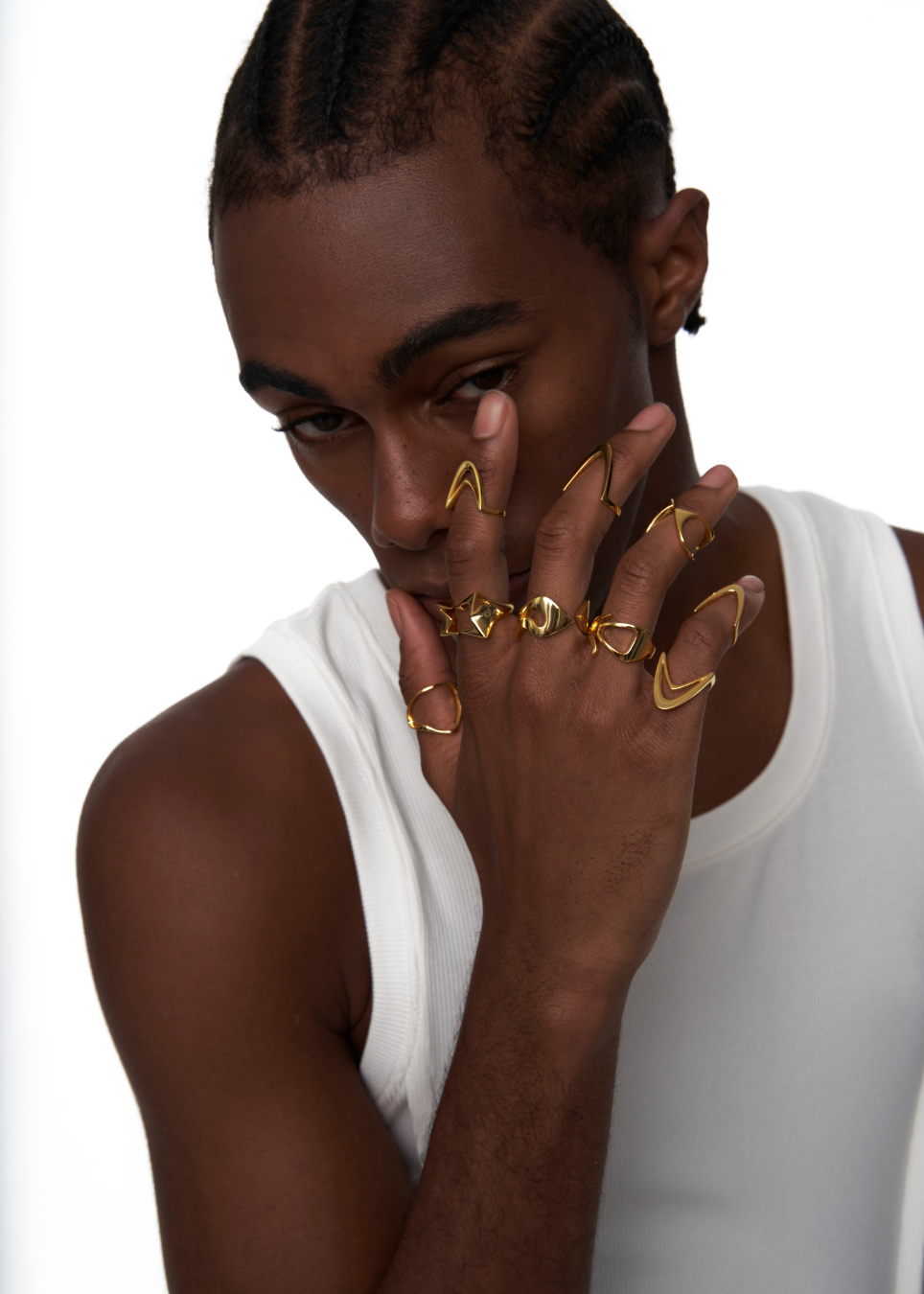 A close-up of a model wearing multiple gold rings on their fingers and hand, styled with a white tank top against a clean white background.