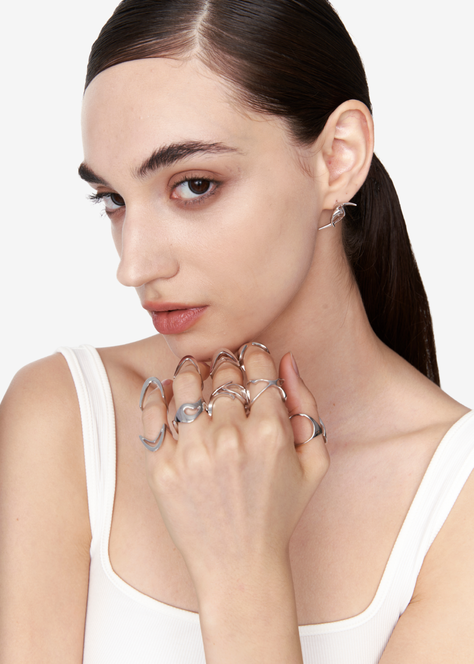 A close-up of a model wearing multiple silver rings on her fingers, including a large circular ring and several slim bands, with a small silver earring visible. She is wearing a white tank top against a clean white background.