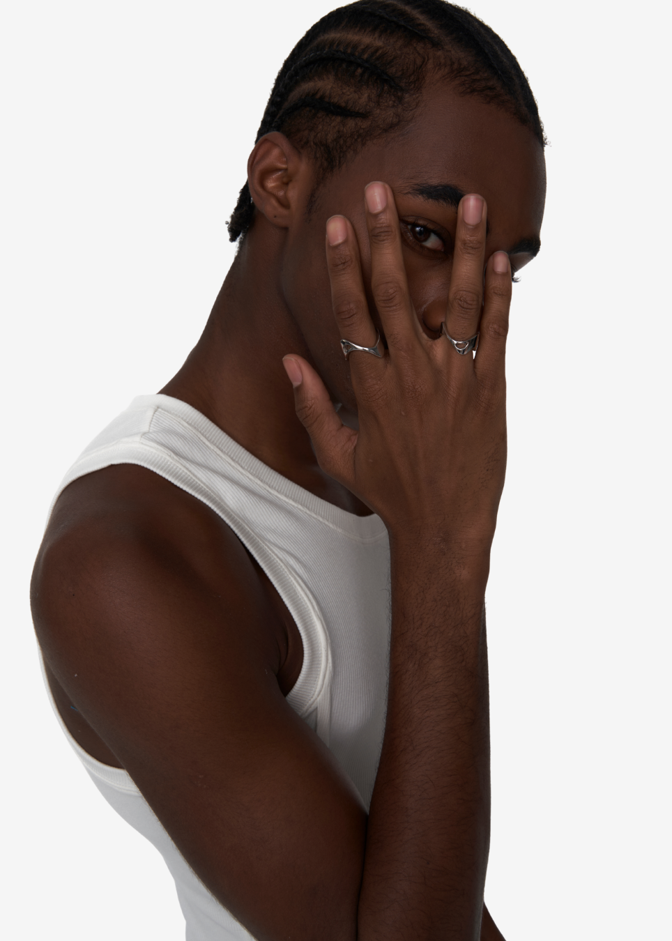 A man wearing a white ribbed tank top and silver rings, posing with his hand over his face against a plain white background.
