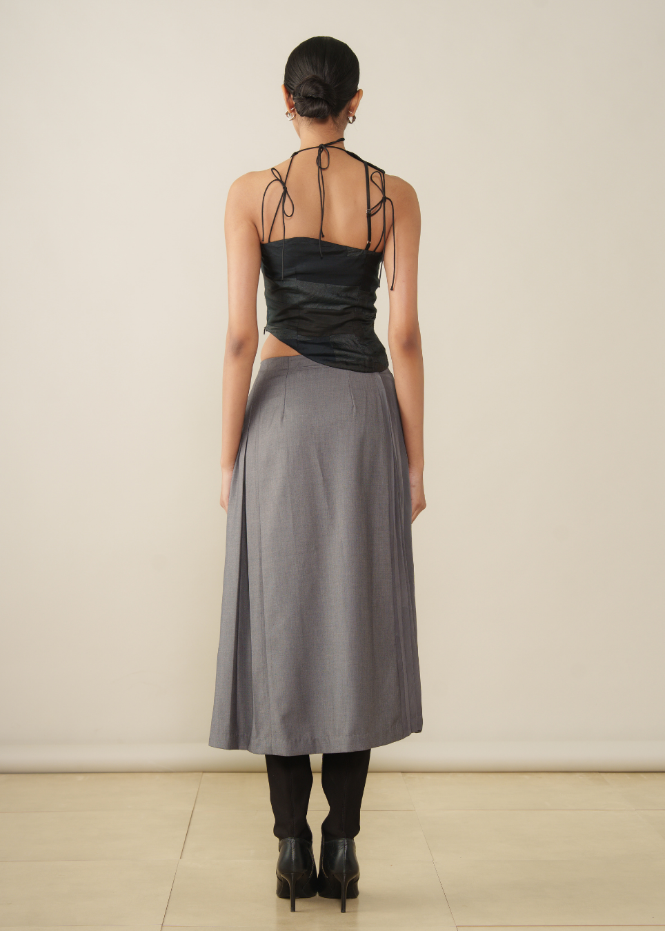 A model shown from the back wearing a black halter-neck top with strappy details and a high-waisted gray midi skirt, paired with black tights and black ankle boots, against a plain beige studio background.