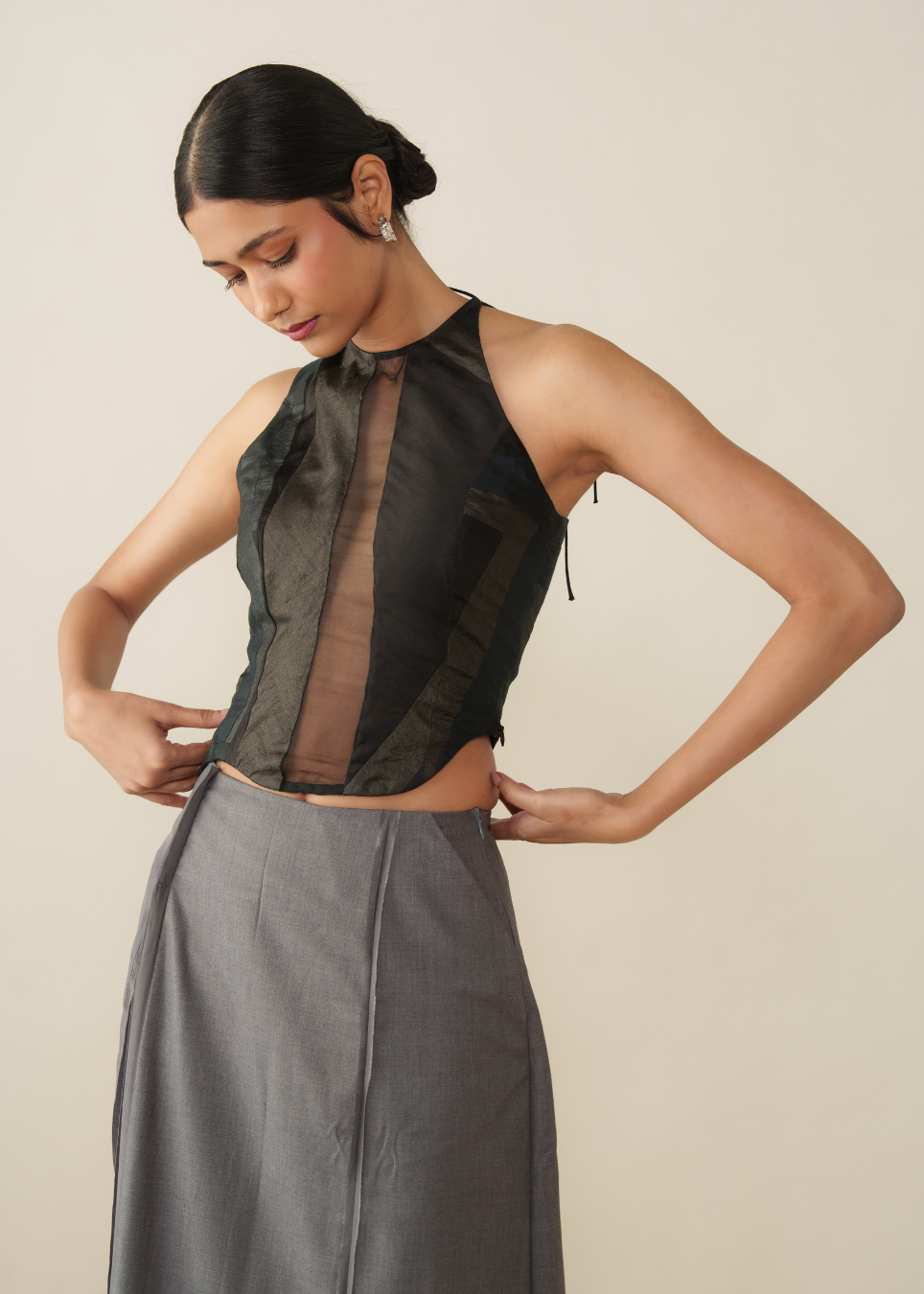 A woman modeling a sleeveless black sheer mesh top with a sheer brown front panel and a high neck, paired with a gray skirt, against a plain beige background.