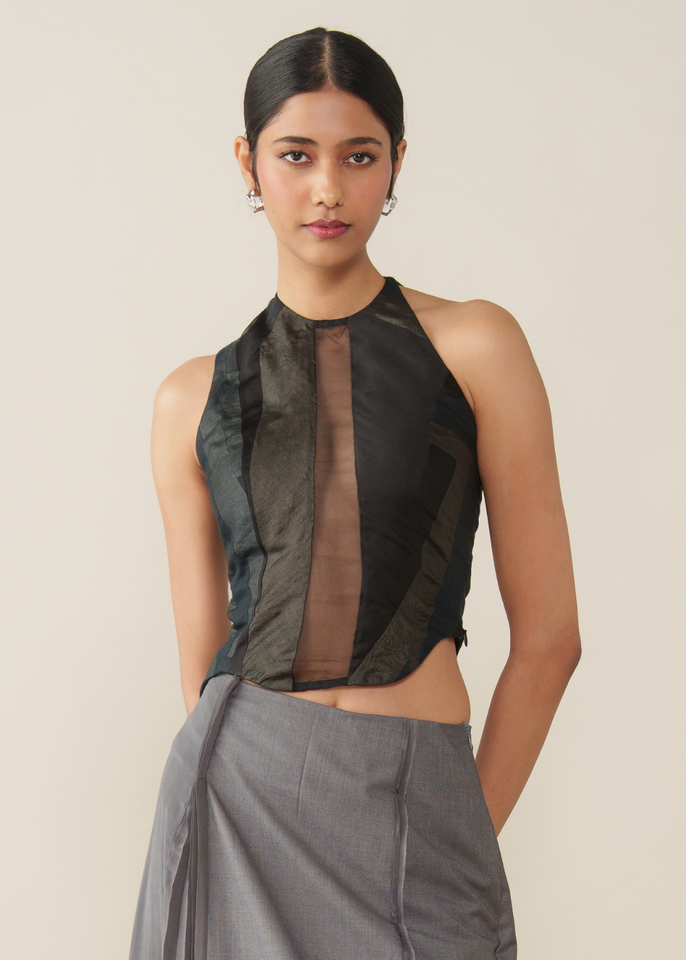 A model wearing a sleeveless high-neck crop top with a mixed texture of black, dark gray, and sheer brown panels, paired with a gray draped skirt, against a solid beige background.