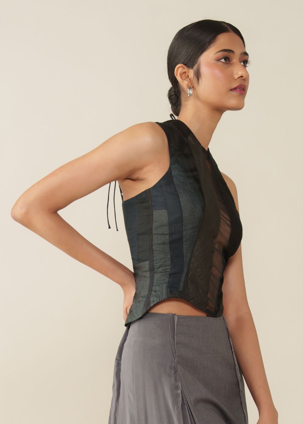 A woman wearing a sleeveless black and dark gray corset-style top with sheer mesh panels and tie details, paired with gray trousers, against a plain beige studio background.
