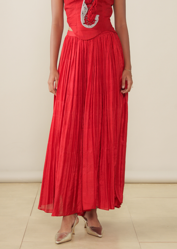 A model wearing a vibrant red pleated maxi skirt paired with a matching red top featuring silver and white embroidery, styled with metallic gold heels against a neutral beige background.