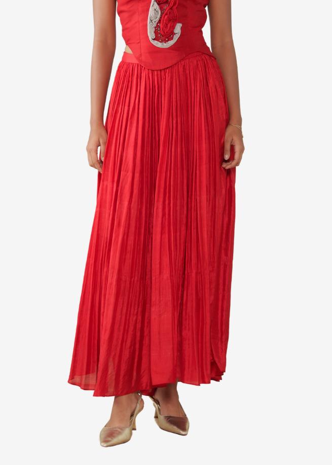 A woman modeling a sleeveless red maxi pleated skirt and a silver paisley embellishment on the bodice, set against a plain light gray background.