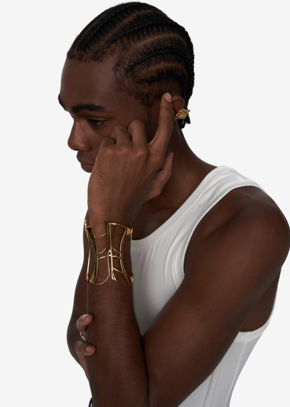A man in a white tank top shown in profile, wearing a wide gold cuff bracelet on his wrist and a matching gold hoop earring in his ear. The background is a clean, solid light gray.