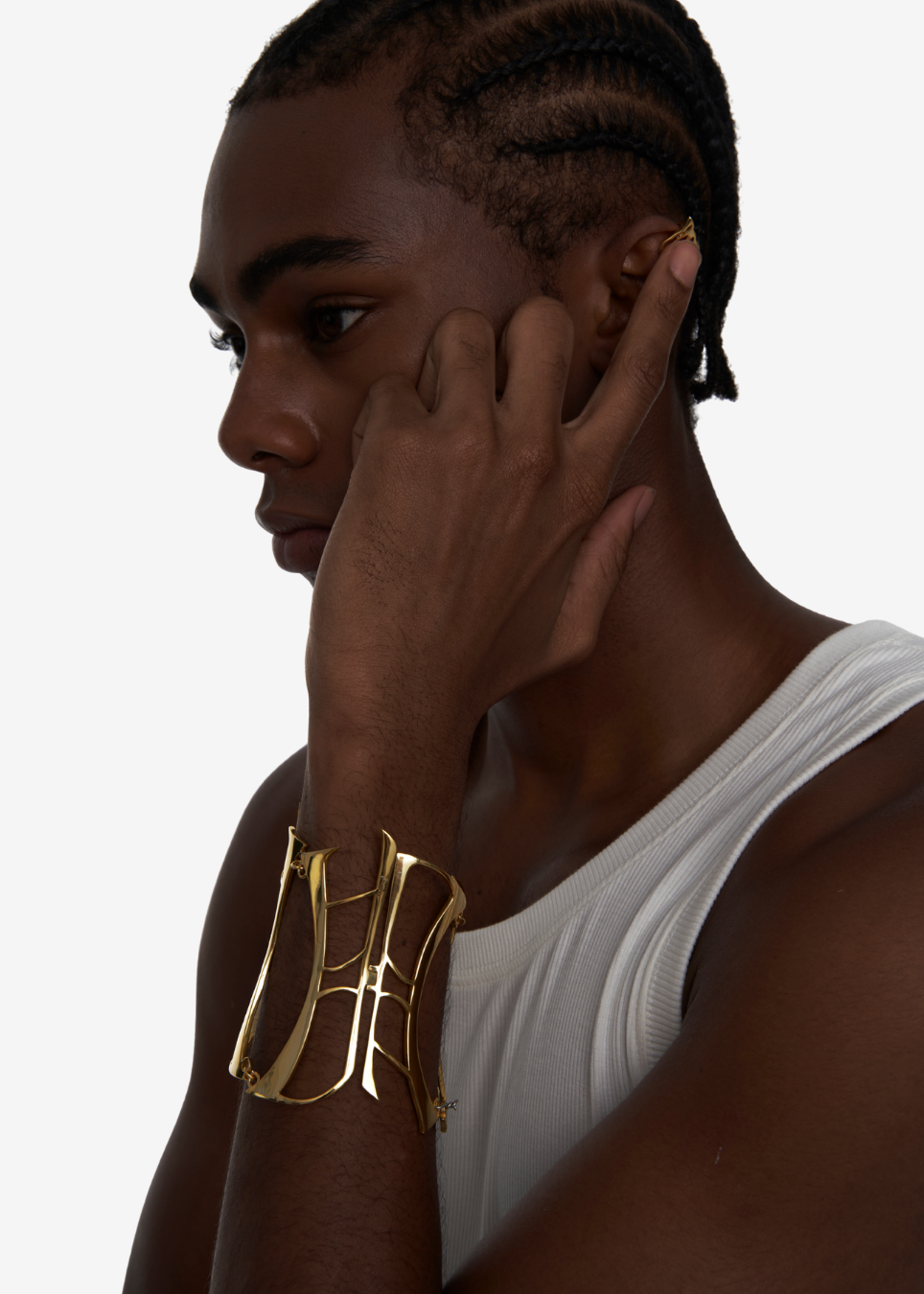 A man in profile wearing a wide, sculptural gold cuff bracelet on his wrist and a matching gold hoop earring in his ear, against a clean white background.