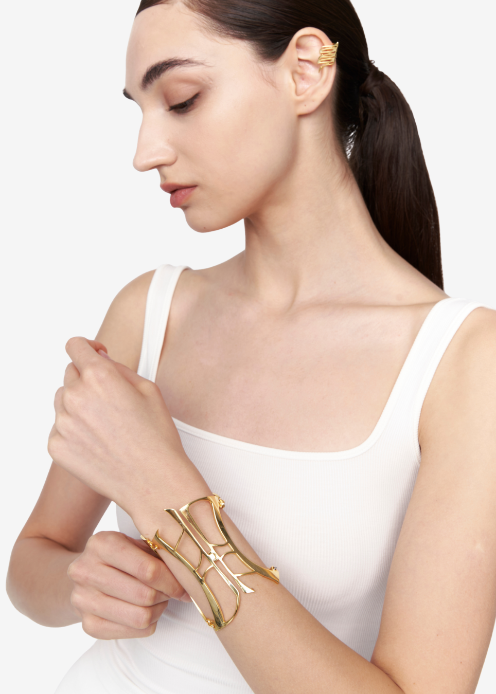 A woman wearing a white ribbed tank top models a gold-toned geometric cuff bracelet on her wrist against a clean white background.