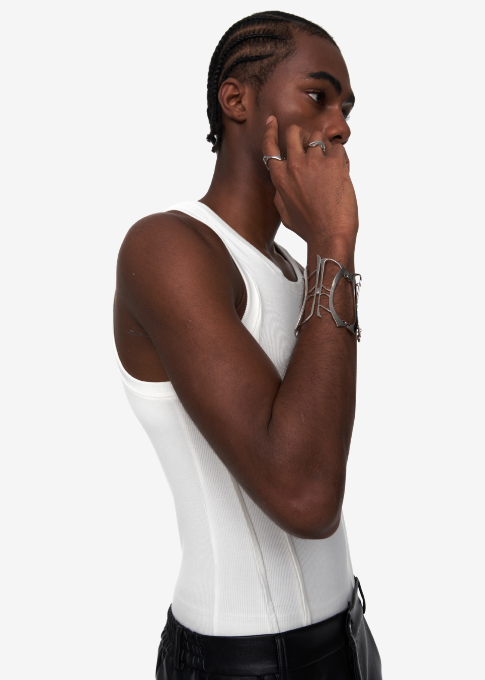 A man in profile wearing a fitted white ribbed tank top and black pants, with silver bracelets and rings, against a plain light gray studio background.