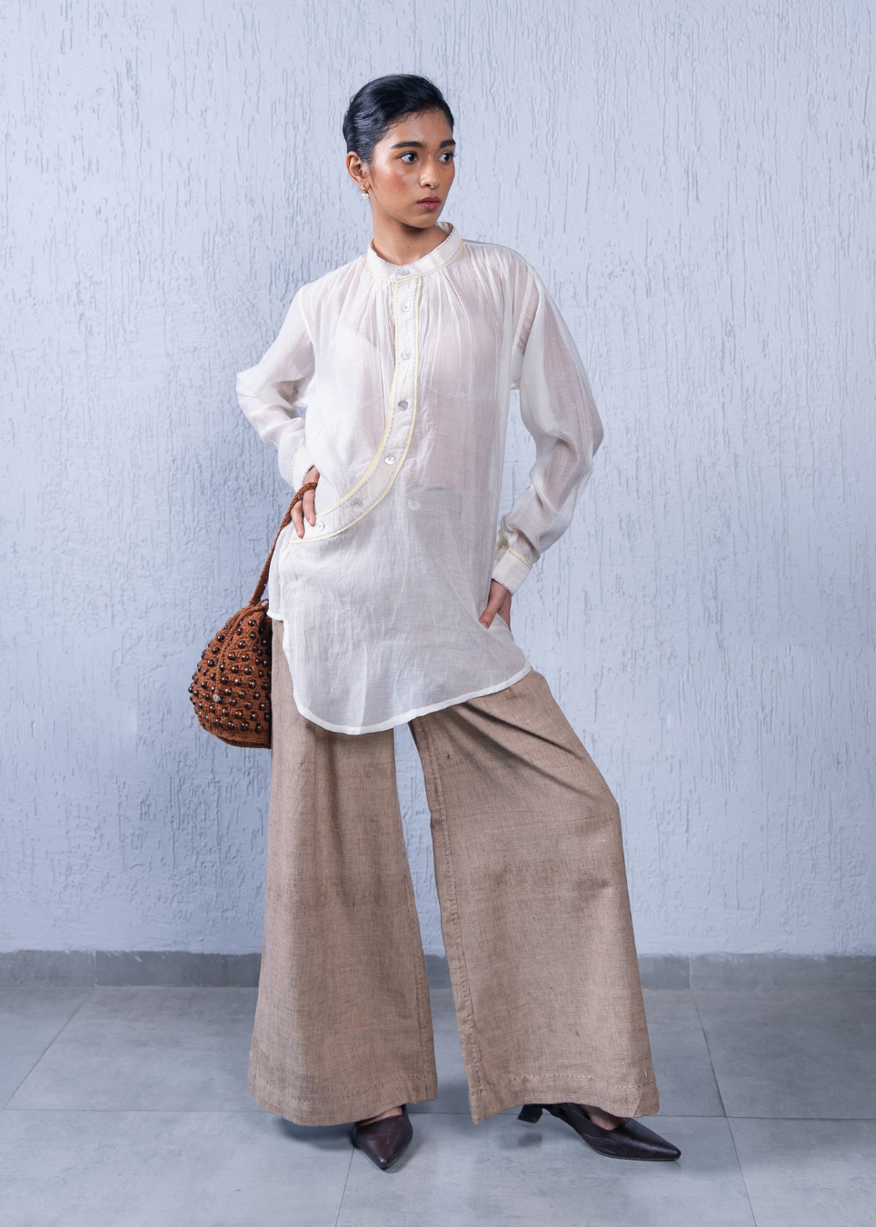 A woman models a white long-sleeve button-up tunic with gold embroidery, paired with wide-leg beige trousers, dark brown shoes, and a brown woven shoulder bag, standing against a textured light gray wall.