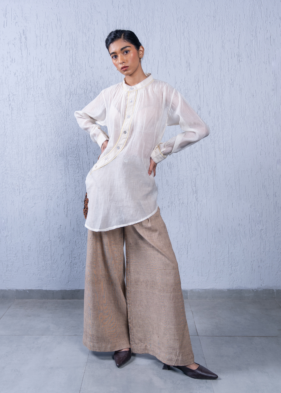 A woman modeling a white long-sleeve button-front tunic top with beige wide-leg trousers, standing against a textured light gray wall.
