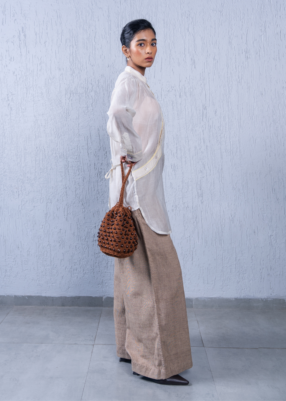A woman stands in profile against a light gray textured wall, wearing a sheer white long-sleeve top, wide-leg beige trousers, and black pointed shoes, while carrying a brown woven bucket bag.