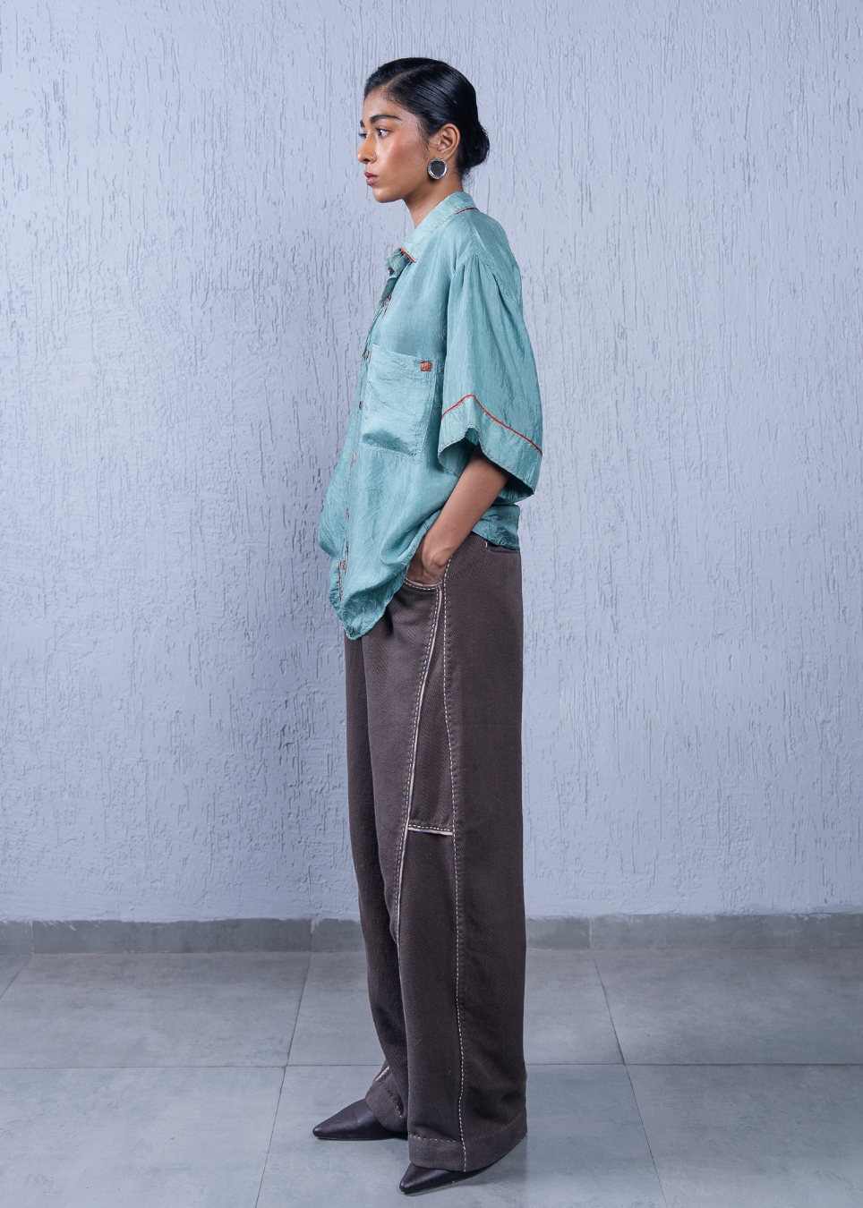 A model stands in profile wearing a light teal oversized button-up shirt with contrast stitching and dark brown wide-leg pants, set against a textured light gray wall and tiled floor.