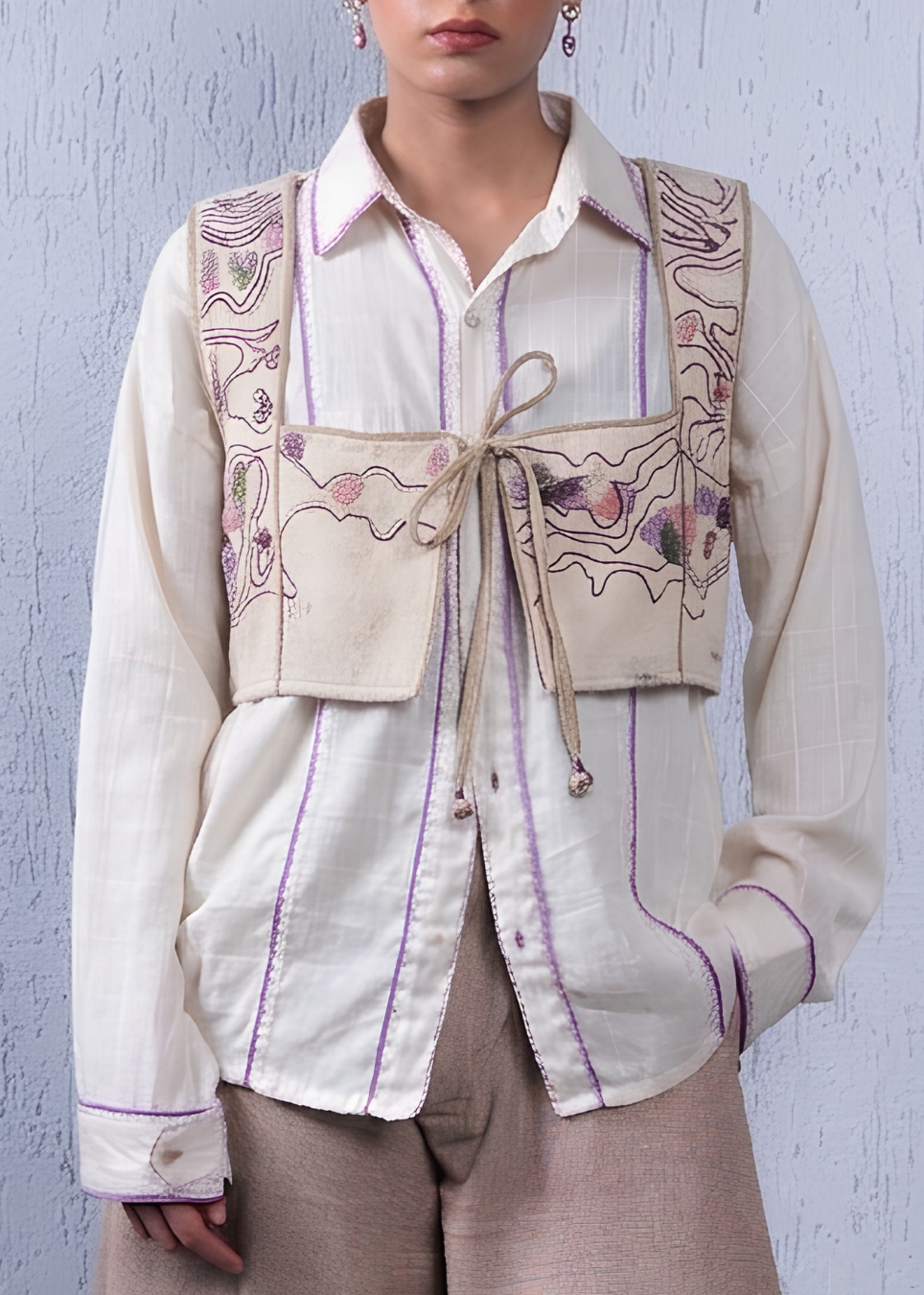 A model wearing a cream-colored long-sleeve button-up shirt with purple piping and a beige vest featuring colorful floral embroidery and a front tie closure, paired with beige trousers against a textured light blue wall.