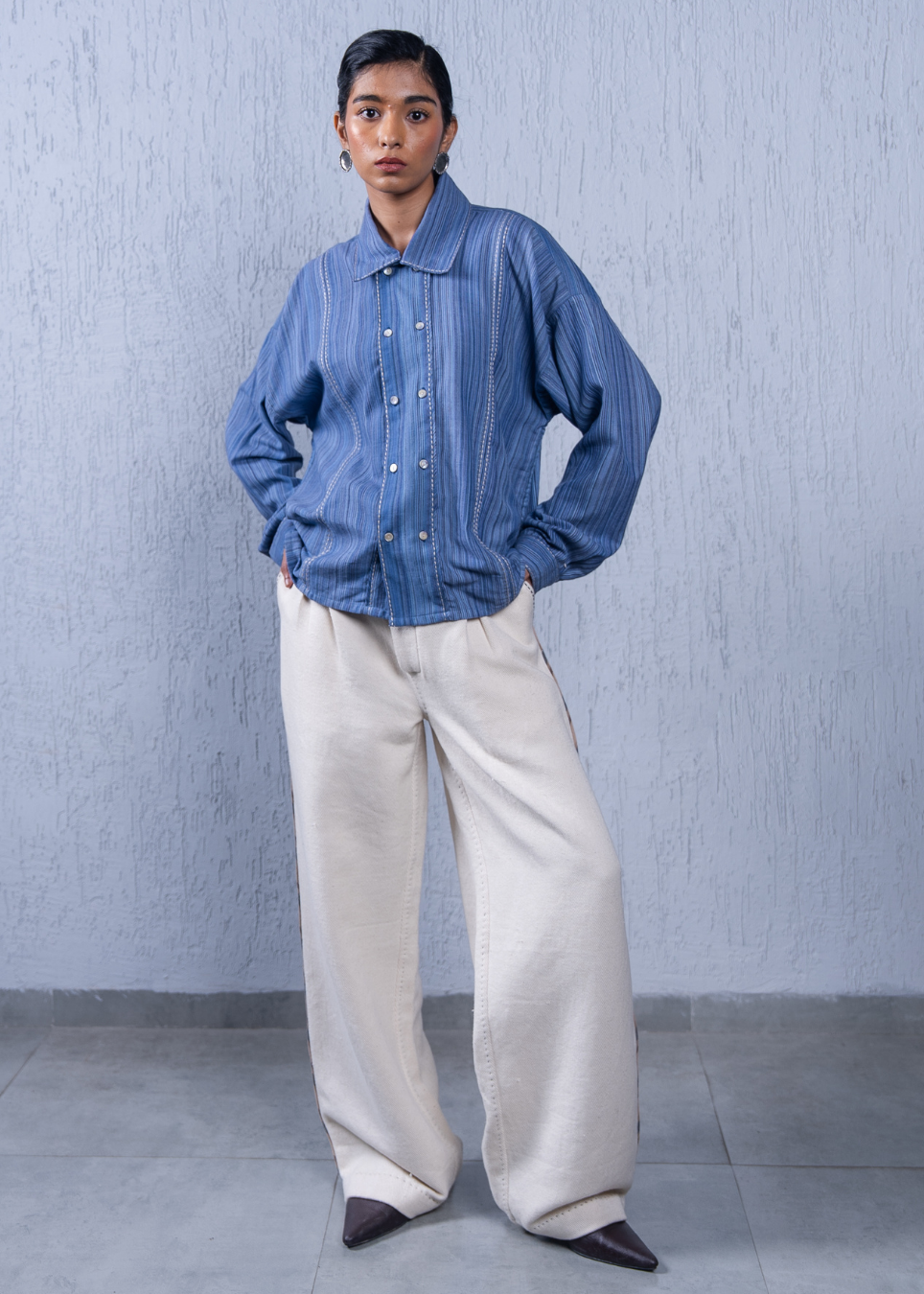 A model stands against a textured light blue-gray wall wearing a blue and white vertically striped button-up shirt and cream wide-leg trousers.