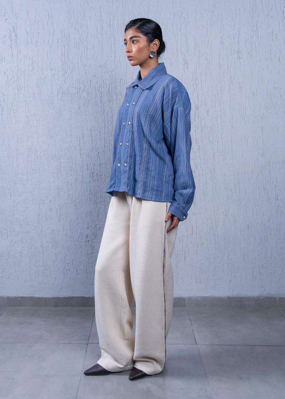 A woman stands in profile wearing a blue vertically striped button-up shirt and cream wide-leg trousers against a textured light gray wall and gray floor.