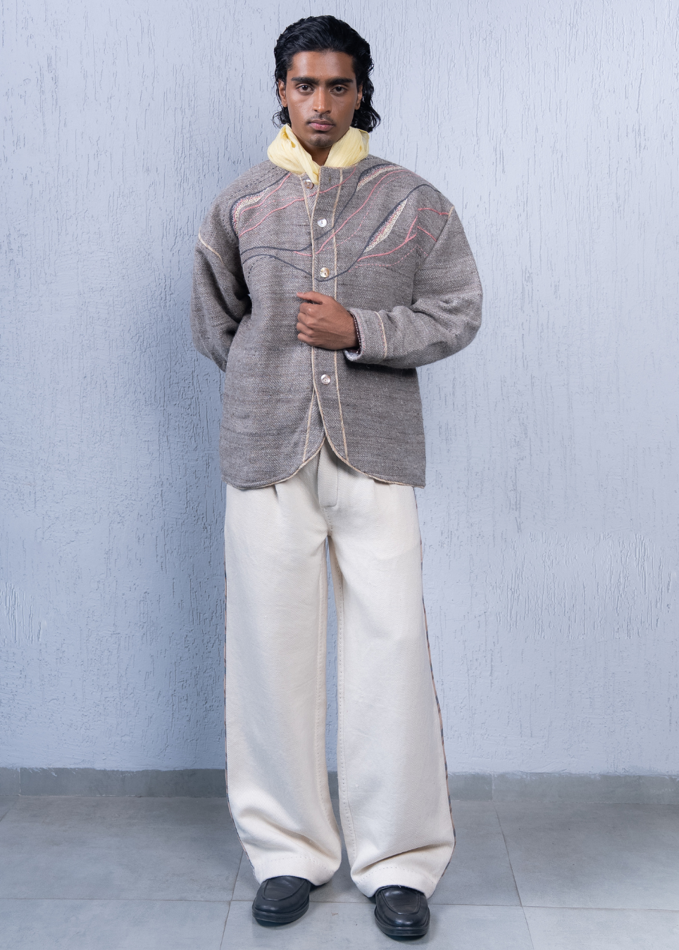 A man modeling a textured gray button-up jacket with a yellow collar, paired with cream wide-leg trousers and black shoes, standing against a light gray textured wall.