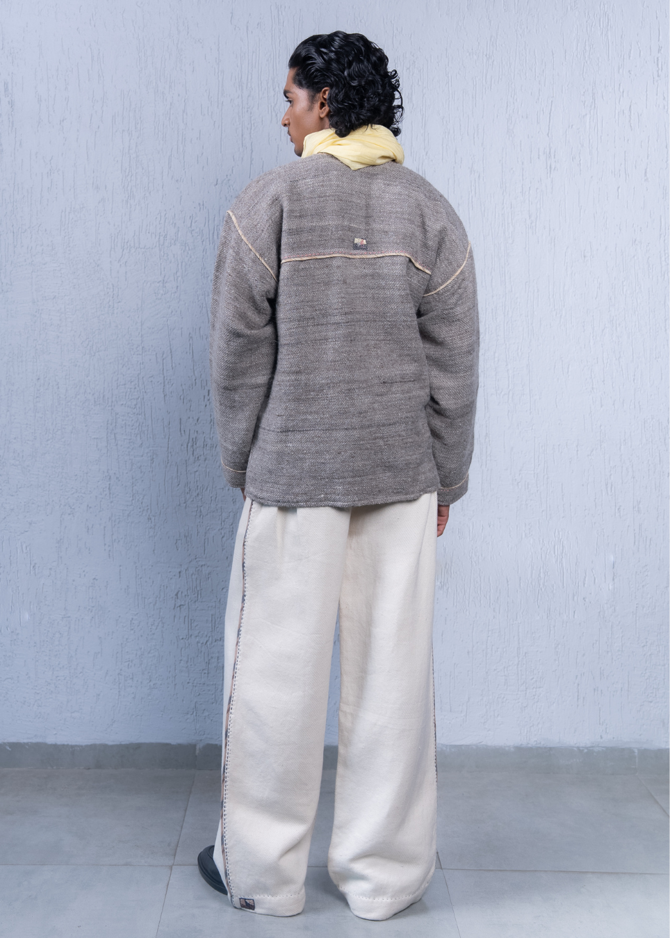 A person shown from the back wearing a textured gray cropped jacket with contrast stitching, cream wide-leg trousers, and a yellow collar layer, standing against a light blue-gray wall.