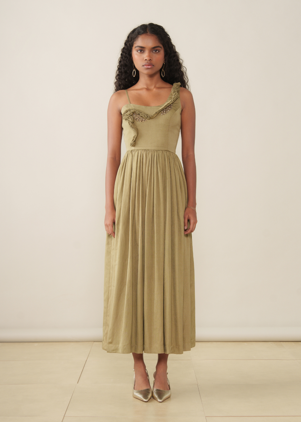 A woman stands against a plain cream wall wearing a long olive-beige dress with a ruffled asymmetrical neckline and a flowing pleated skirt, paired with pointed metallic heels.