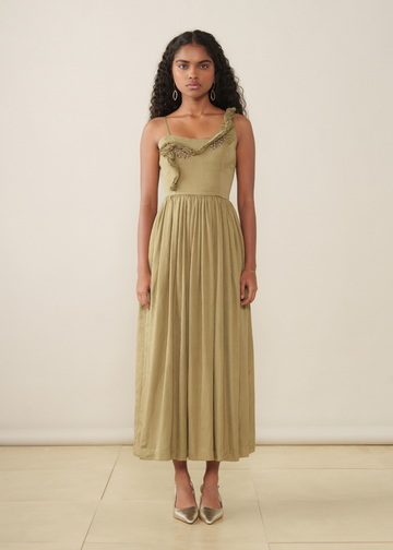 A woman stands against a plain cream wall wearing a long olive-beige dress with a ruffled asymmetrical neckline and a flowing pleated skirt, paired with pointed metallic heels.