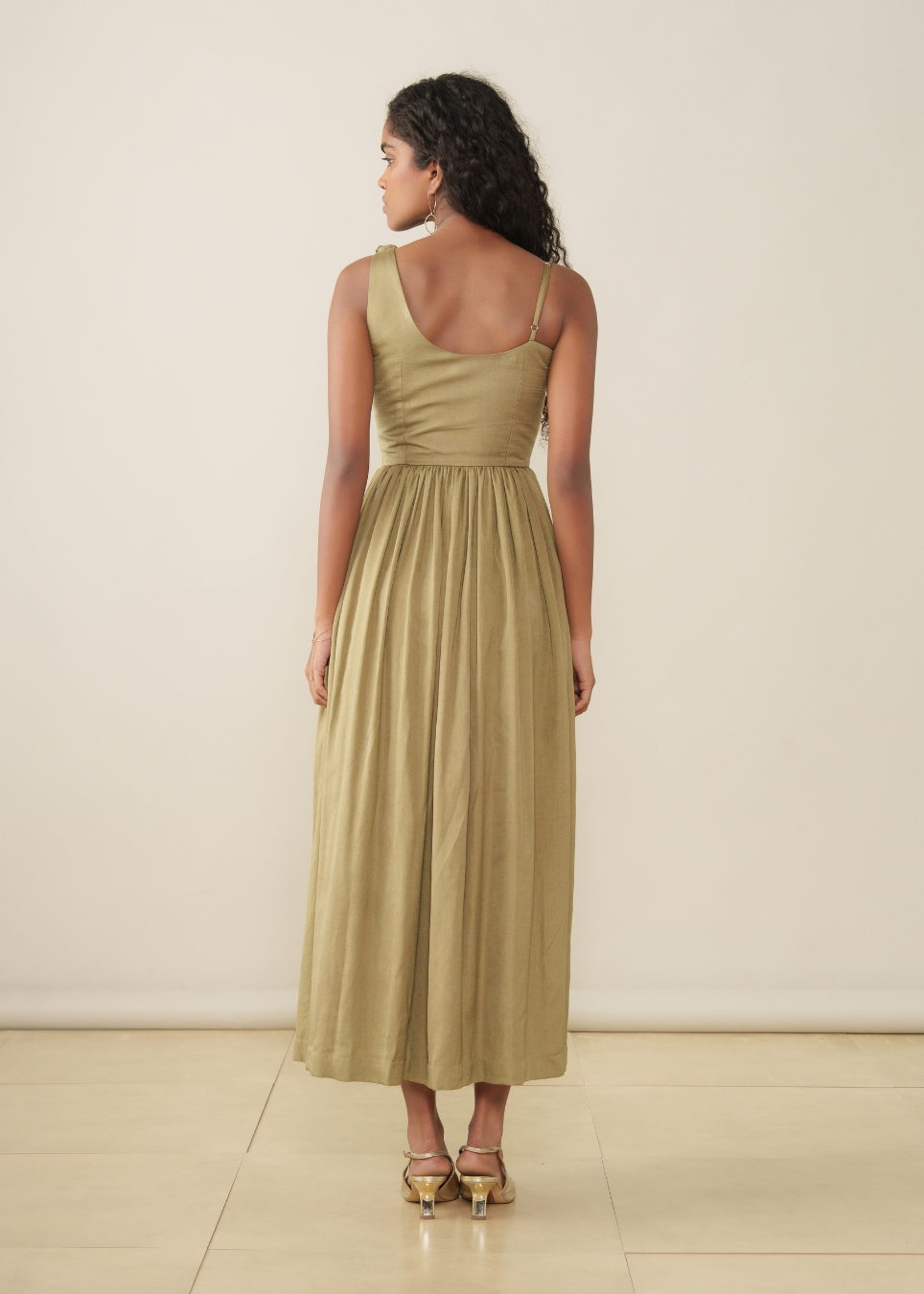 A woman stands with her back to the camera wearing a sleeveless, scoop-neck maxi dress in a muted olive-beige color, paired with gold heeled sandals, against a plain cream studio background.