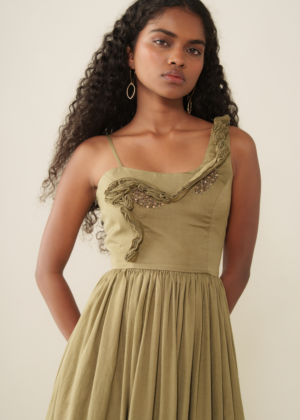 A woman modeling a one-shoulder olive green dress with a ruffled embellished bodice and a pleated skirt, set against a plain light beige background.