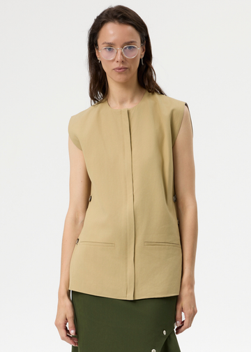 A woman wearing a sleeveless beige vest with two front flap pockets, paired with an olive green skirt, against a plain light gray background.