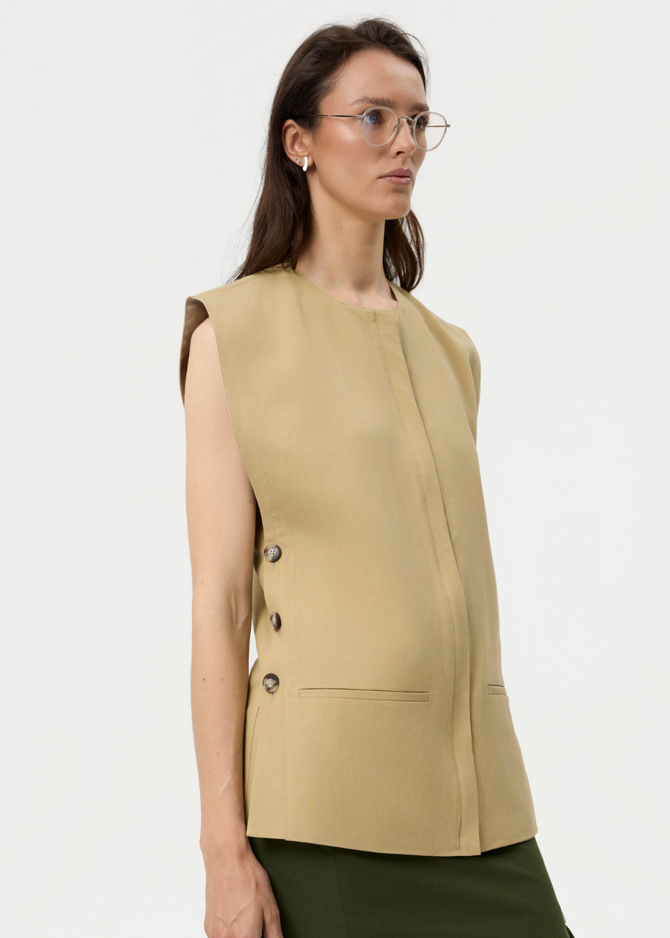 A woman wearing a sleeveless beige vest with three dark buttons on the side and dark green bottoms, photographed against a clean white studio background.