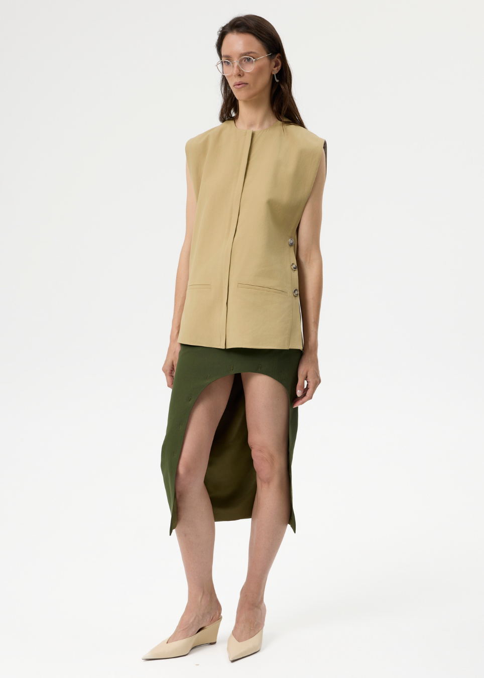 A model stands against a plain white studio background wearing a sleeveless beige vest over an olive green asymmetrical skirt, paired with beige pointed-toe heels.