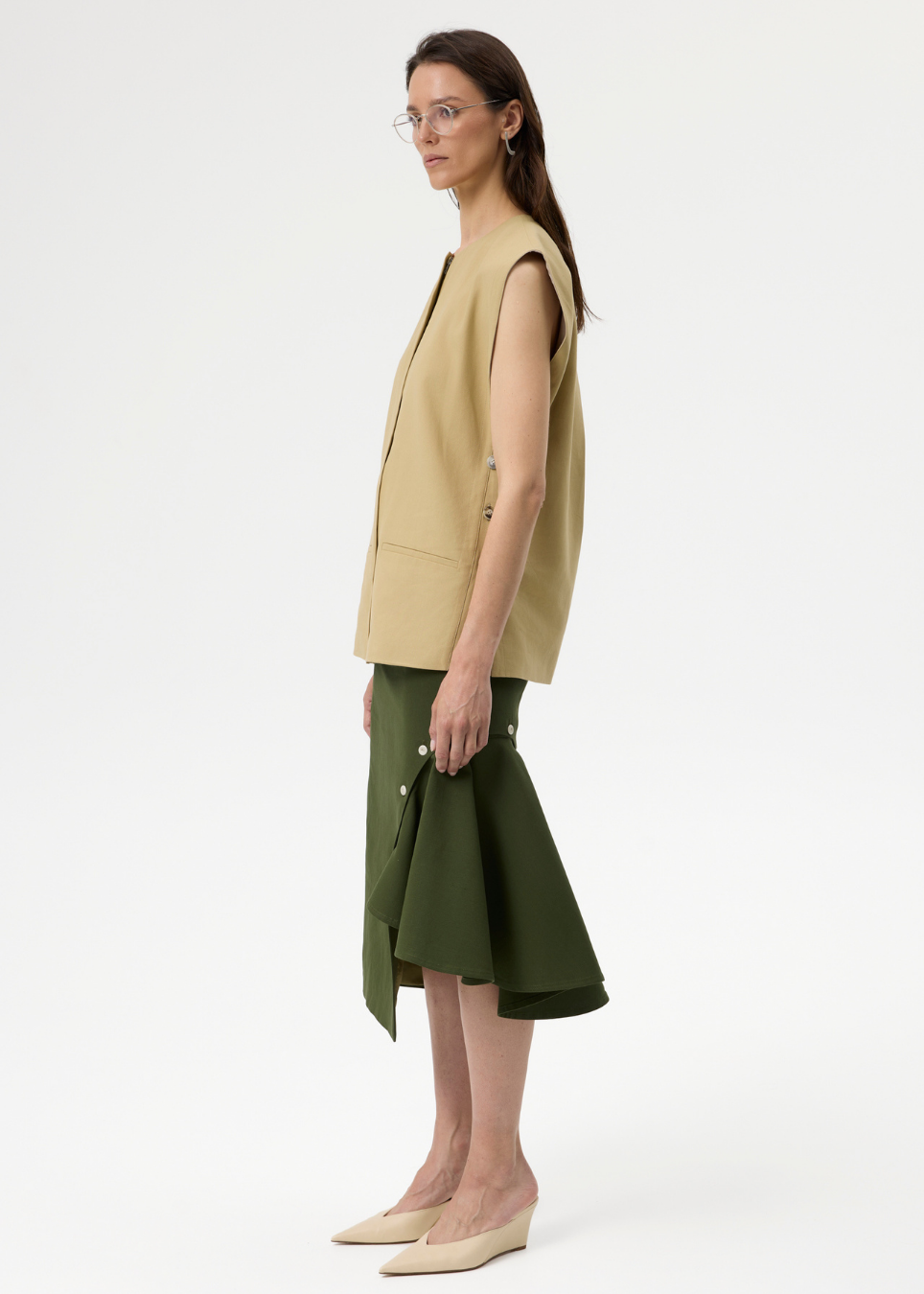 A model is shown in profile wearing a sleeveless beige vest, an olive green midi skirt, and beige wedge pumps against a plain white studio background.