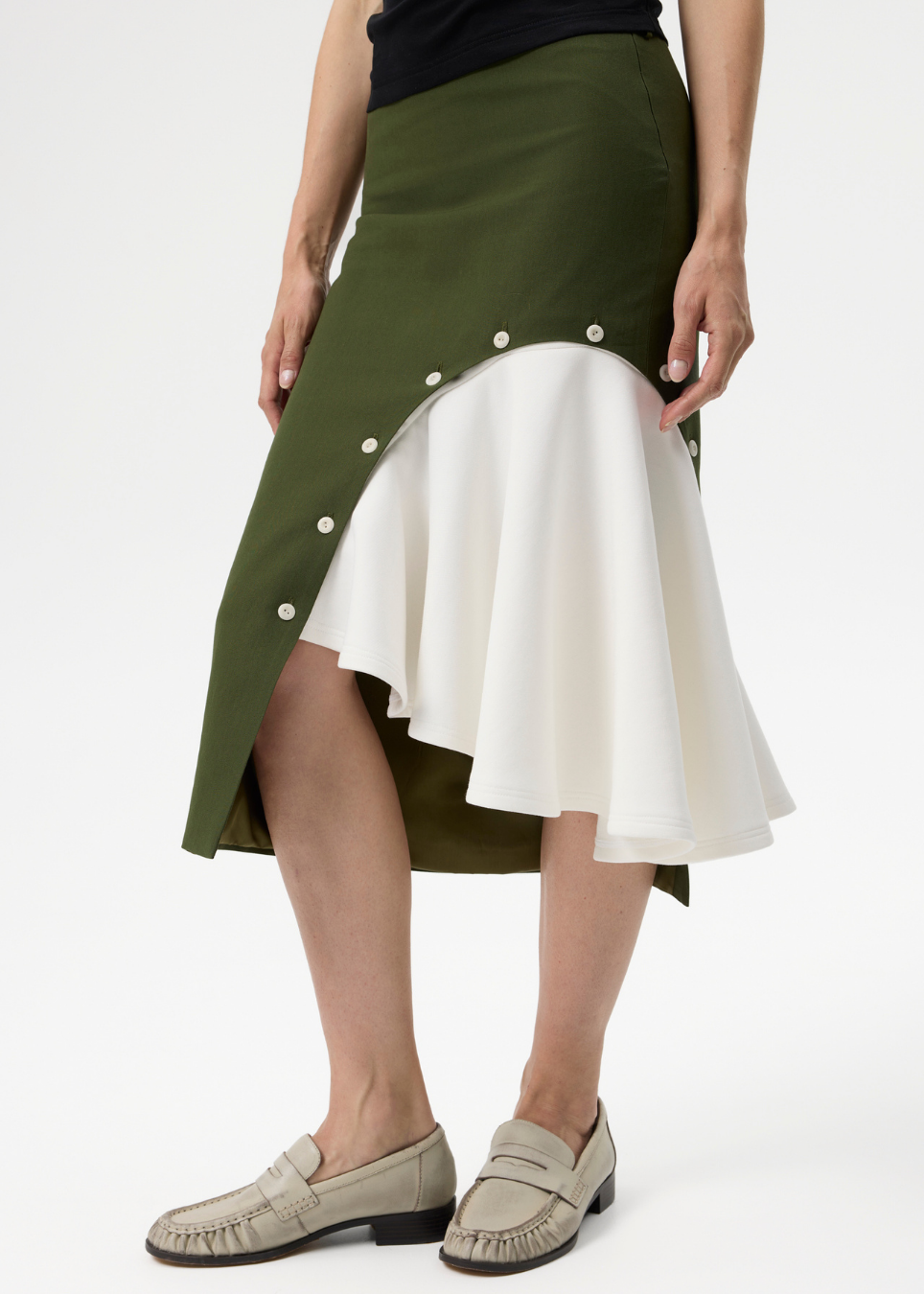 A model wearing a high-low asymmetrical skirt with an olive green outer layer adorned with white buttons and a layered white flared inner layer, paired with beige loafers against a plain white studio background.