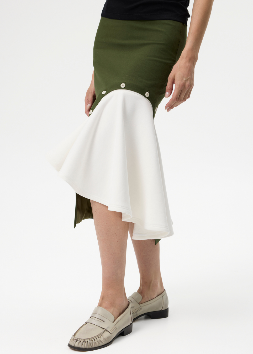 A model wearing a high-low asymmetrical skirt with an olive green upper section and a flowing white flared lower section, paired with beige loafers on a white studio background.