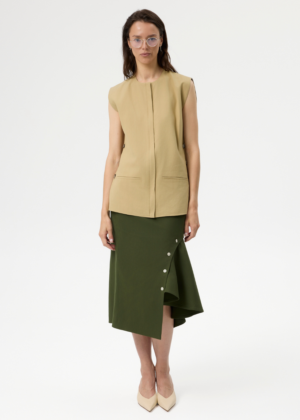 A model stands against a plain light gray studio background wearing a sleeveless beige vest and an olive green asymmetrical skirt with side buttons, paired with beige heels.