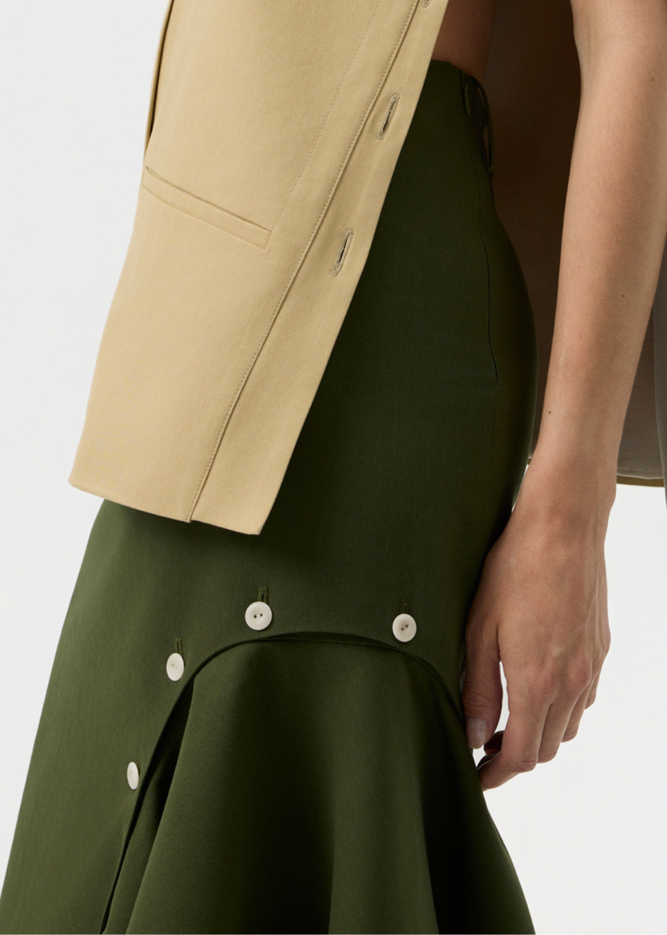 Close-up of a model wearing an olive green skirt with decorative buttons and a beige top, against a plain light background.